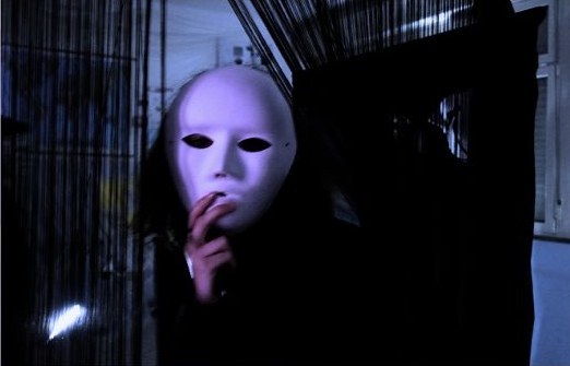 Una persona con una maschera bianca dietro una tenda di fili scuri, durante il museo
